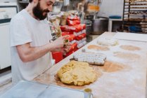 Man in white t-shirt putting fresh dough into cups while making pastry in kitchen of bakery — Stock Photo