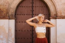 Jovem mulher atraente em roupas elegantes braços dobráveis e fechar os olhos enquanto está de pé contra o edifício antigo — Fotografia de Stock
