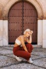 Приваблива молода жінка в стильному вбранні, присідаючи, дивлячись на стародавню будівлю з бурхливими воротами на вулиці старого міста — стокове фото