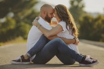 Side view of happy couple in matching casual clothes embracing and looking at each other while sitting on countryside road — Stock Photo