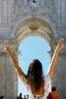 Back view of woman in sunglasses with hands up showing v sign while standing beside majestic arch in city — Stock Photo