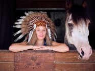 Delighted woman in authentic Indian feather hat leaning on wooden fence in stable with horse on ranch and looking at camera — Stock Photo