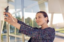 Femme entrepreneure gaie dans une veste élégante souriante et utilisant un smartphone pour prendre selfie à l'extérieur du bâtiment moderne — Photo de stock
