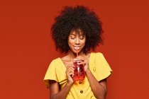 Verträumte Afroamerikanerin mit lockigem Haar hält ein rotes Glas mit Stroh in der Hand und genießt das Getränk auf rotem Hintergrund — Stockfoto