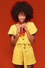 Dreamy African American woman with curly hair holding red jar with straw and enjoying beverage on red background — Stock Photo