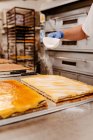 Cocinero anónimo derramando azúcar en polvo de la pala encima de la torta sabrosa mientras trabaja en la cocina de la panadería - foto de stock