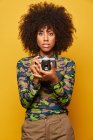 Професійна жінка-фотограф з зачіскою в голові тримає фотокамеру, стоячи на жовтому тлі. — стокове фото