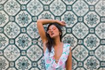 Мирная роскошная женщина в модном наряде и блестящих солнцезащитных очках, опирающаяся на мозаичную экзотическую стену с закрытыми глазами на живописной улице — стоковое фото