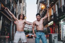 Gritando hombres multiétnicos quitándose las camisas y saludando por encima de la cabeza mientras están de pie en la calle urbana de verano - foto de stock