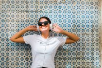 Bela mulher em roupa casual e fones de ouvido com as mãos para cima ao lado parede de mosaico azul de construção na rua da cidade — Fotografia de Stock