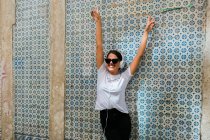 Hermosa mujer en traje casual y auriculares con las manos arriba junto a la pared de mosaico azul del edificio en la calle de la ciudad - foto de stock
