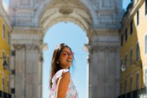 Young cheerful woman in sunglasses standing beside majestic arch in city street in Lisbon, Portugal — Stock Photo