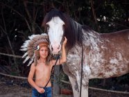 Delighted kid in Indian feather war bonnet caressing horse on ranch and looking at camera — Stock Photo