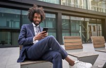 Gioioso uomo africano americano in abito seduto fuori e messaggistica — Foto stock