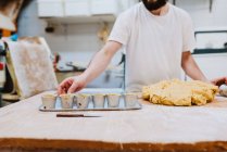 Бородатый мужчина в белой футболке кладет свежее тесто в чашки, пока печет тесто на кухне пекарни — стоковое фото