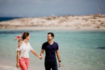 Hombre y mujer sonrientes caminando a lo largo de la orilla del mar mientras se toman de la mano y se miran con amor sobre un fondo borroso - foto de stock