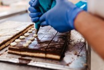 Nahaufnahme anonymer Mitarbeiter in Handschuhen, der Sahne auf frischem Schokoladenkuchen auf Tablett in Bäckerei drückt — Stockfoto