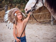 Criança encantada em penas indiana boné de guerra acariciando cavalo no rancho — Fotografia de Stock