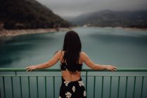 From behind slim brunette in open back dress leaning on handrail of bridge across still lake at scenic mountain valley — Stock Photo