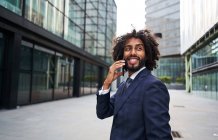 Alegre hombre afroamericano en traje formal parado afuera y hablando en un teléfono inteligente mientras mira hacia otro lado - foto de stock
