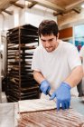 Mann in Uniform und Handschuhen schneidet mit Messer süßen frischen Kuchen auf Tisch in Bäckerei — Stockfoto