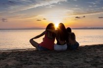 Rückansicht von brünierten Freundinnen, die sich bei Sonnenuntergang im Sommer im Gegenlicht am Strand umarmen und entspannen — Stockfoto