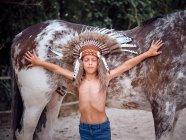 Thoughtful child in Indian feather hat with closed eyes leaning on stallion with spread hands on ranch — Stock Photo