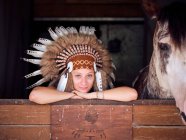 Mulher encantada em chapéu de pena indiana autêntica apoiada em cerca de madeira no estábulo com cavalo no rancho e olhando para a câmera — Fotografia de Stock