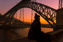 Vista posteriore del turista donna che guarda lontano mentre si siede sul bordo del terrapieno della città vicino al ponte durante il tramonto a Oporto, Portogallo — Foto stock