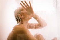 Vista lateral de la mujer desnuda tocando el vidrio mientras está sentada en el suelo y tomando la ducha en casa - foto de stock
