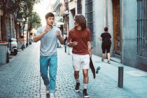 Hombres multiétnicos interesados despreocupados en ropa casual con gestos de longboard y hablar mientras pasean por la calle de la ciudad - foto de stock