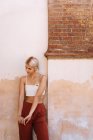 Giovane donna in top alla moda e pantaloni guardando giù mentre in piedi contro muro edificio squallido sulla strada della città antica — Foto stock