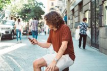 Fröhlicher, lässiger Mann, der auf einem Geländer an der Stadtstraße sitzt, lacht und mit dem Smartphone spricht — Stockfoto