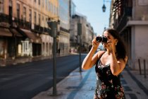 Femme confiante en robe d'été prenant des photos avec caméra alors qu'elle se tenait debout sur la rue ensoleillée pittoresque de Lisbonne, Portugal — Photo de stock