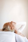 Side view of beautiful blond woman closing eyes and leaning on comfortable mattress against white wall at home — Stock Photo