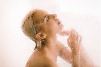 Vue latérale de la jeune femme prenant une douche derrière cloison transparente humide dans la salle de bain — Photo de stock