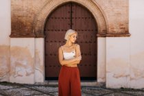 Attraktive junge Frau in stylischem Outfit faltet die Arme und schließt die Augen, während sie vor einem antiken Gebäude mit schäbigem Tor steht — Stockfoto