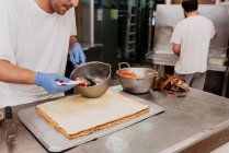 Хлібопекарський працівник в латексних рукавичках, що розкладає солодке варення на свіжій булочці над кухонною стійкою — стокове фото