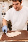 Hombre de la cosecha en guante de látex y uniforme decorando delicioso pastel con remolinos de crema blanca mientras se trabaja en la panadería - foto de stock