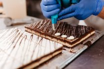 Анонимный работник крупным планом в перчатках, сжимающий крем поверх свежих шоколадных тортов на подносе в пекарне — стоковое фото