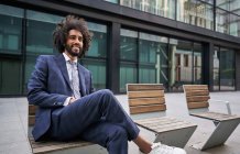 Оптимистичный и веселый афроамериканский предприниматель с бородой сидит на скамейке со скрещенными ногами и смотрит в сторону — стоковое фото