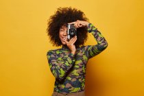 Fotógrafa profesional y alegre que toma fotos con una elegante cámara fotográfica mientras se encuentra en un fondo amarillo. - foto de stock