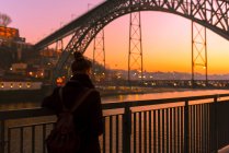 Vue arrière du touriste femelle debout près du remblai de la ville près du pont regardant loin pendant le coucher du soleil à Porto, Portugal — Photo de stock
