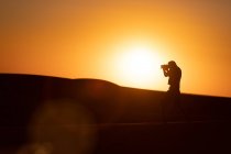 Silhouette inreconnaissable d'un voyageur photographiant des dunes debout sur du sable dans un désert étonnant — Photo de stock