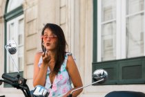 Beautiful woman in sunglasses putting on bright lipstick while looking at motorbike mirror on street — Stock Photo