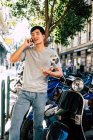 Позитивный случайный азиат говорит по смартфону и жестикулирует, стоя на мотоцикле на солнечной улице — стоковое фото