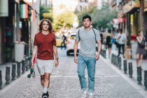 Carefree interested multiethnic men in casual clothes with longboard gesturing and talking while strolling along city street — Stock Photo