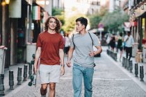 Carefree interested multiethnic men in casual clothes with longboard gesturing and talking while strolling along city street — Stock Photo