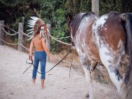 Back view of kid wearing feather Indian war bonnet and walking shirtless on sandy farm, leading horse behind — Stock Photo
