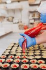 Anonymous confectioner in latex gloves squeezing tasty sweet jelly from bag into small pastry cases on blurred background of bakery kitchen — Stock Photo
